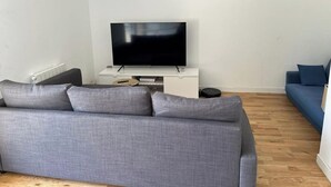 A modern living room with a grey sofa, a flat-screen TV on a white stand, and a blue sofa against a white wall. - Apartment near Paris (Noisy-le-Grand)