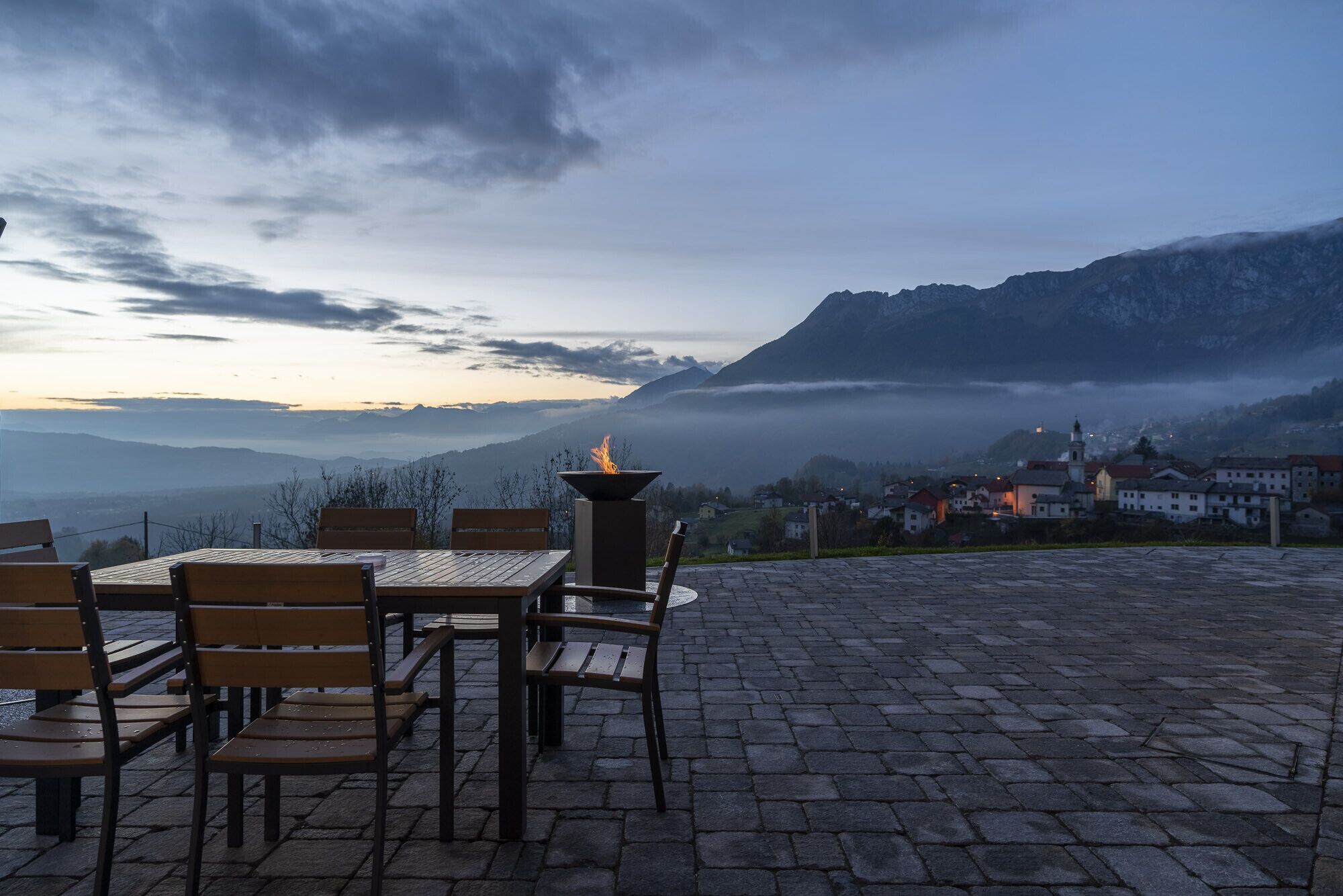 A terrace with stone flooring and wooden chairs, overlooking a mountainous landscape with a village below.