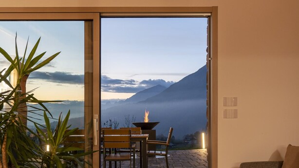 A modern interior with a wooden floor, a sofa, a dining table with chairs, and a large sliding glass door leading to an outdoor patio with a fire feature and a view of mountains. - Casera Cal De Mez Sot - Wellness Chalet (Chies d´Alpago)
