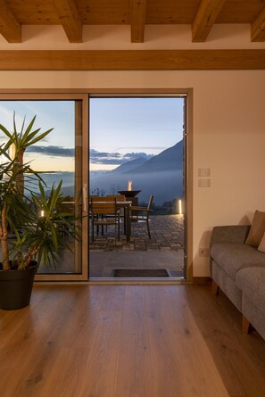 Ein modernes Interieur mit Holzboden, einer Couch, einem Esstisch mit Stühlen und einem großen Schiebeglastür, die auf eine Terrasse mit Feuerstelle und Bergblick führt.