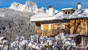 A wooden cabin with a snow-covered roof, a wooden deck, and a mountain backdrop. - CHALET INES - Appartamento 1 (Vodo Cadore)