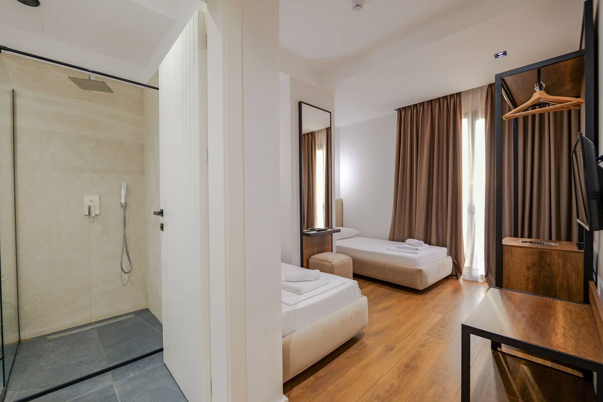 A hotel room with a shower, two beds, a wooden floor, a TV, and a mirror.