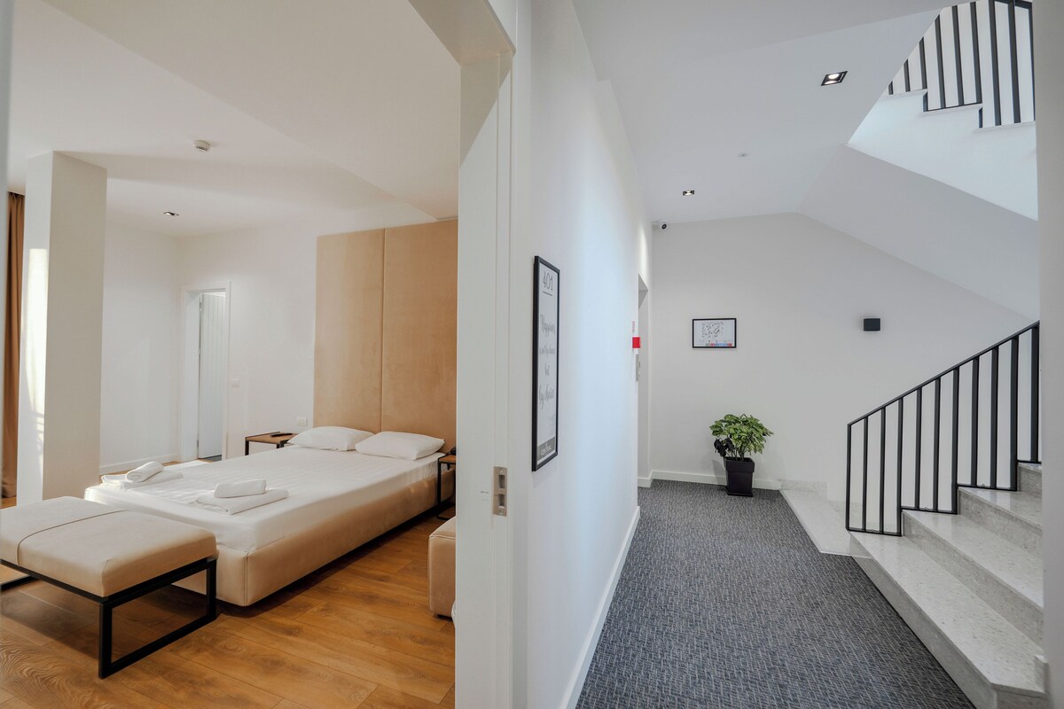 A modern hotel room with a bed, a bench, and a small table. There is a staircase with a railing leading to an upper level.