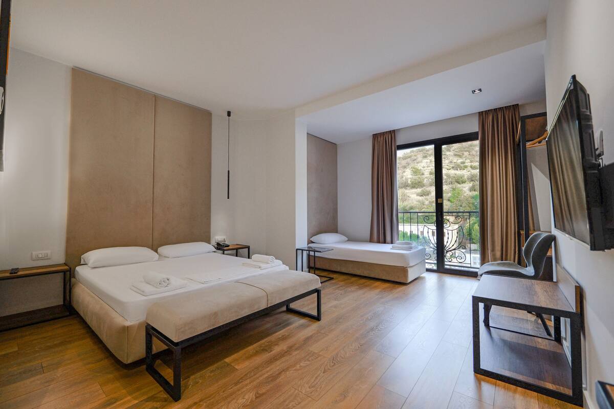 A modern hotel room with a large bed, a sofa, a small table, and a balcony with curtains.