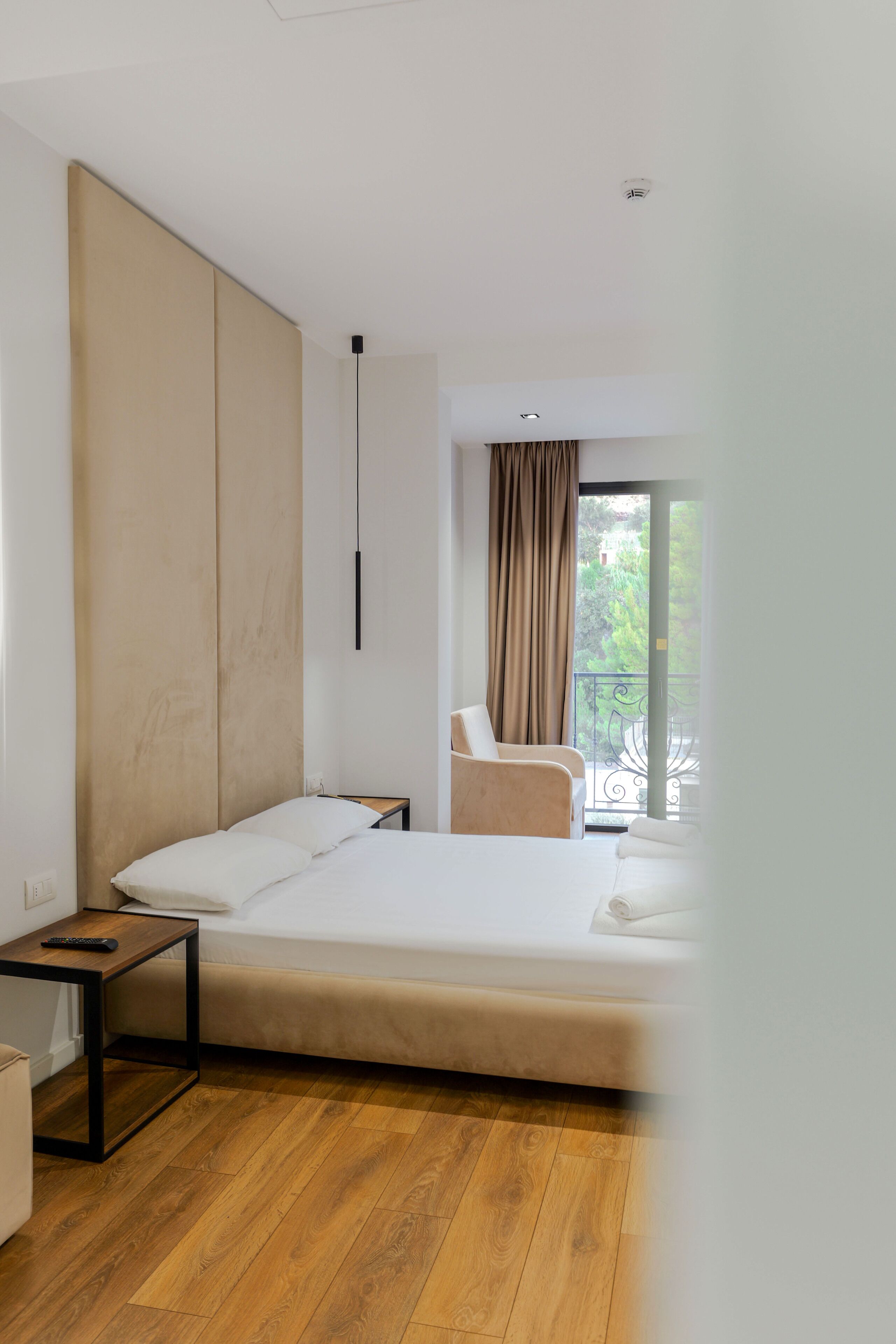 A modern hotel room with a bed, a small table, a chair, and a view of greenery outside.