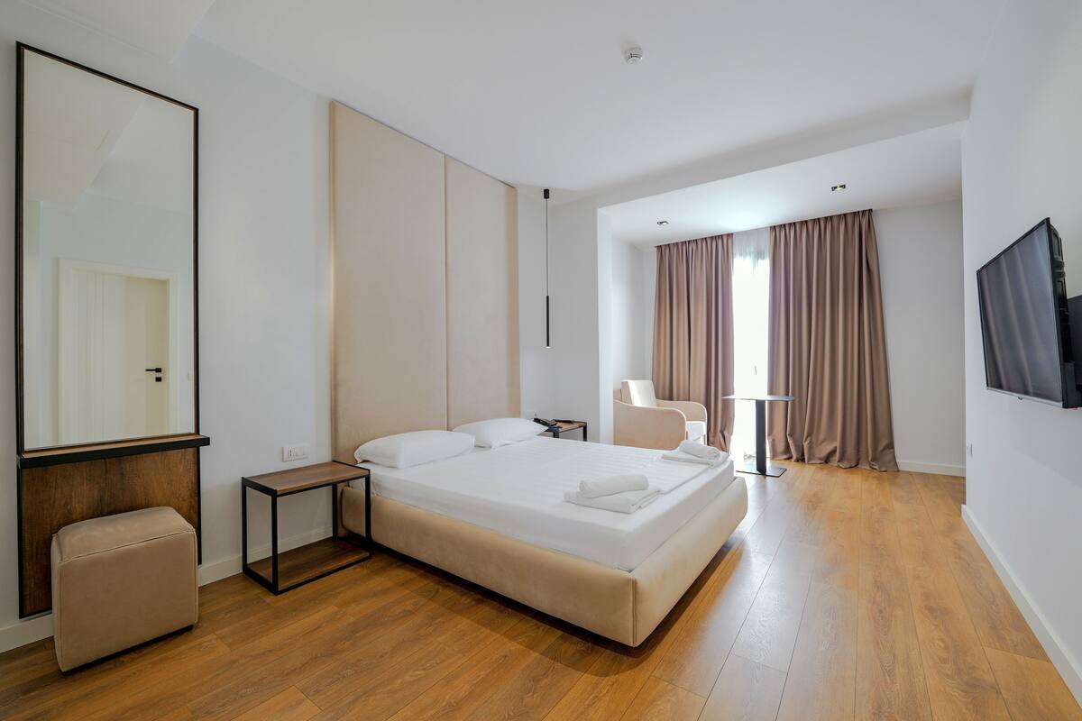 A modern hotel room with a large bed, a bedside table, a chair, a TV, and a large mirror.