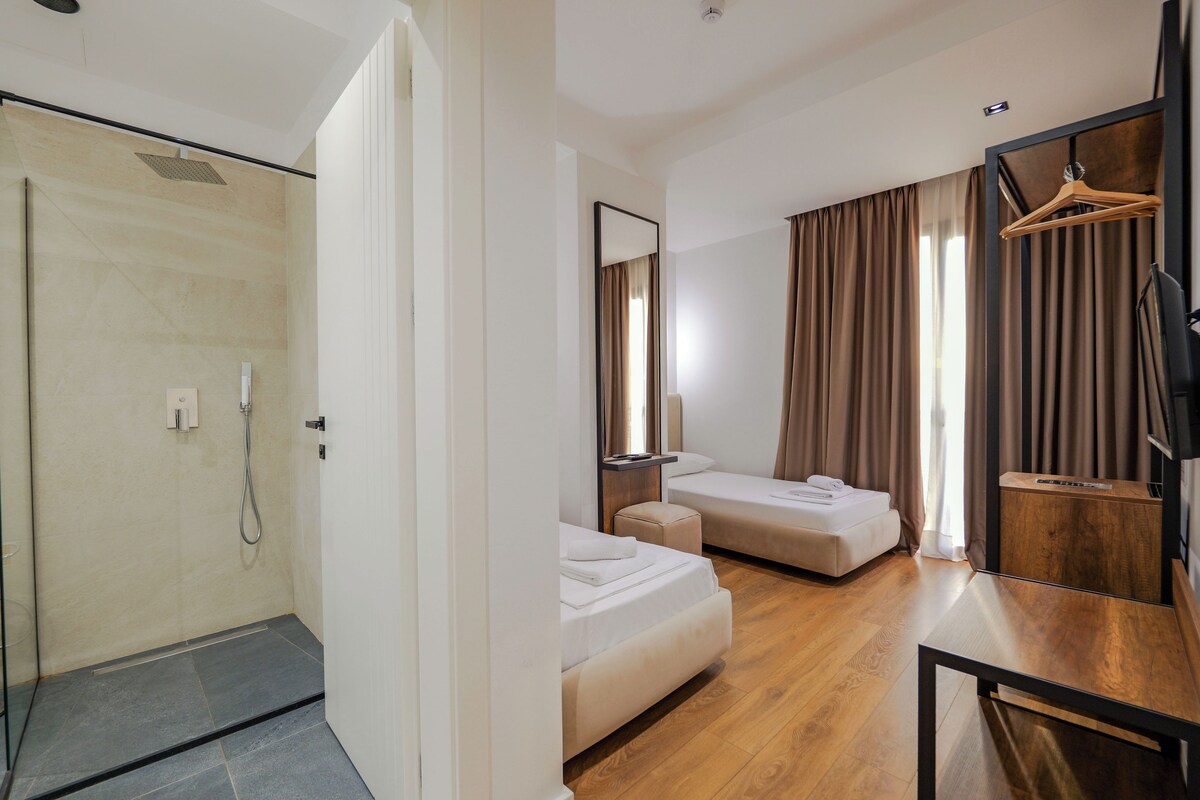 Habitación de hotel con ducha, dos camas, suelo de madera, televisor y espejo.