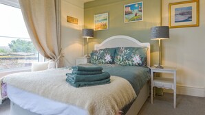 A neatly made bed with folded towels, a white headboard, and a nightstand with a lamp. There are two paintings on the wall and a window with curtains. - Valley View (Teignmouth)