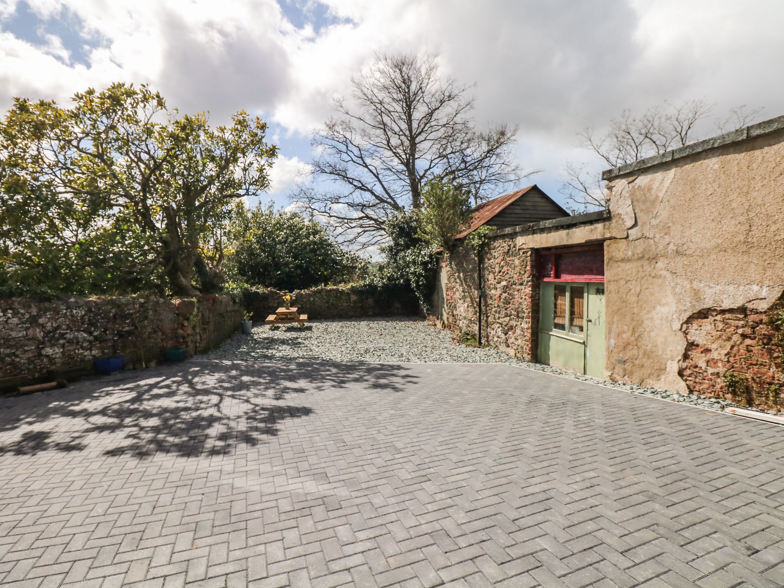 Un chemin pavé mène à un bâtiment rustique en pierre, doté d’une porte rouge, et est entouré d’arbres et d’une zone recouverte de gravier.
