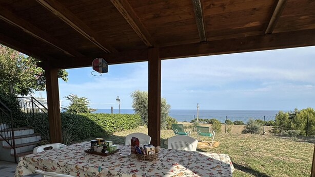 Une terrasse extérieure avec une table dressée pour deux personnes, entourée de chaises en plastique blanches, sous une pergola en bois offrant une vue sur la mer.