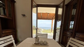 Un coin repas avec une table dressée pour quatre personnes, une machine à café et une vue sur la mer par une porte ouverte.