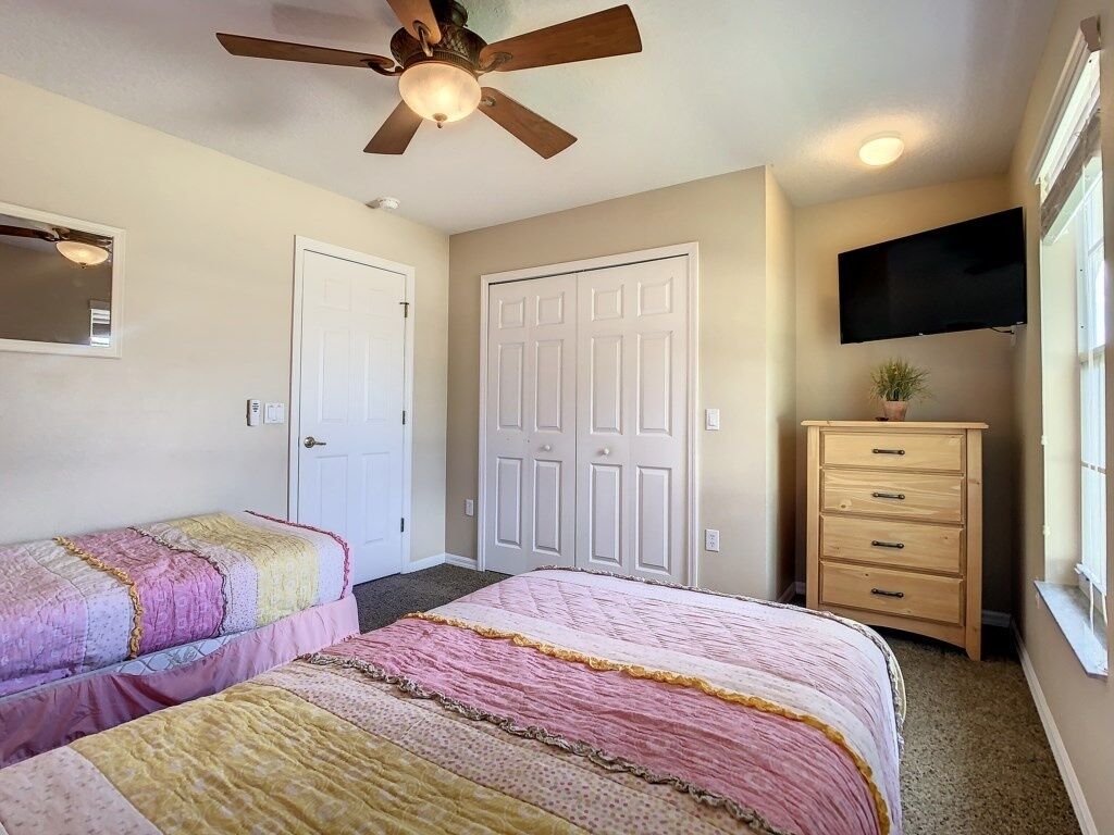 Une chambre avec deux lits, un ventilateur de plafond, une télévision, une commode et une porte.