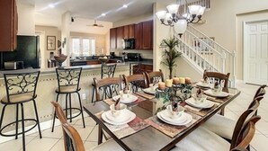 A spacious dining area with a long table set for six, surrounded by wooden chairs. A chandelier hangs above, and a staircase is visible in the background leading to another level.