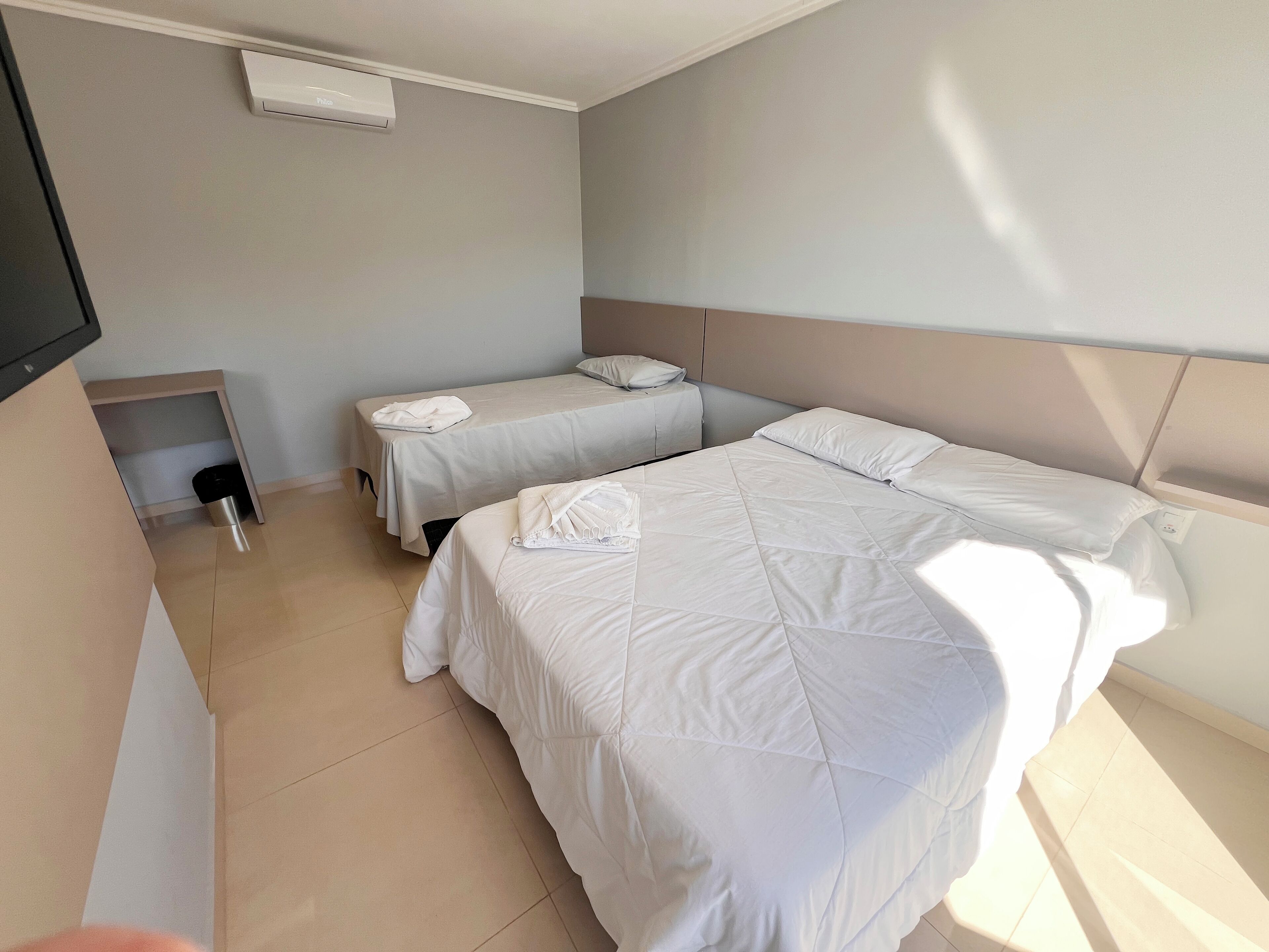 A hotel room with two beds, a small table, an air conditioning unit, and a TV mounted on the wall.