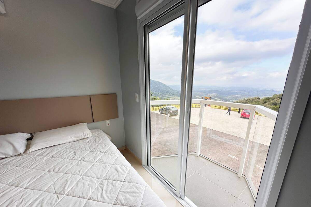 Et soveværelse med en stor seng, en sengegavl og en skydedør, der fører til en balkon med udsigt over en parkeringsplads og et fjerntliggende landskab.