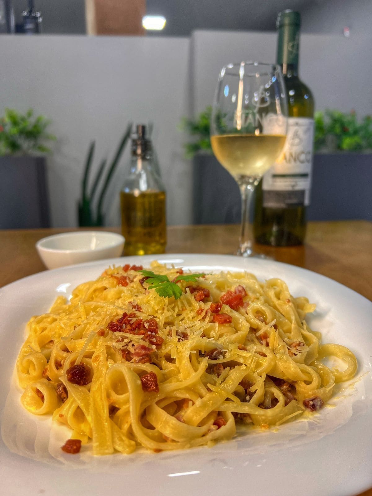 Une assiette de pâtes agrémentée d’une garniture, un verre de vin blanc et deux bouteilles d’huile d’olive sur une table.