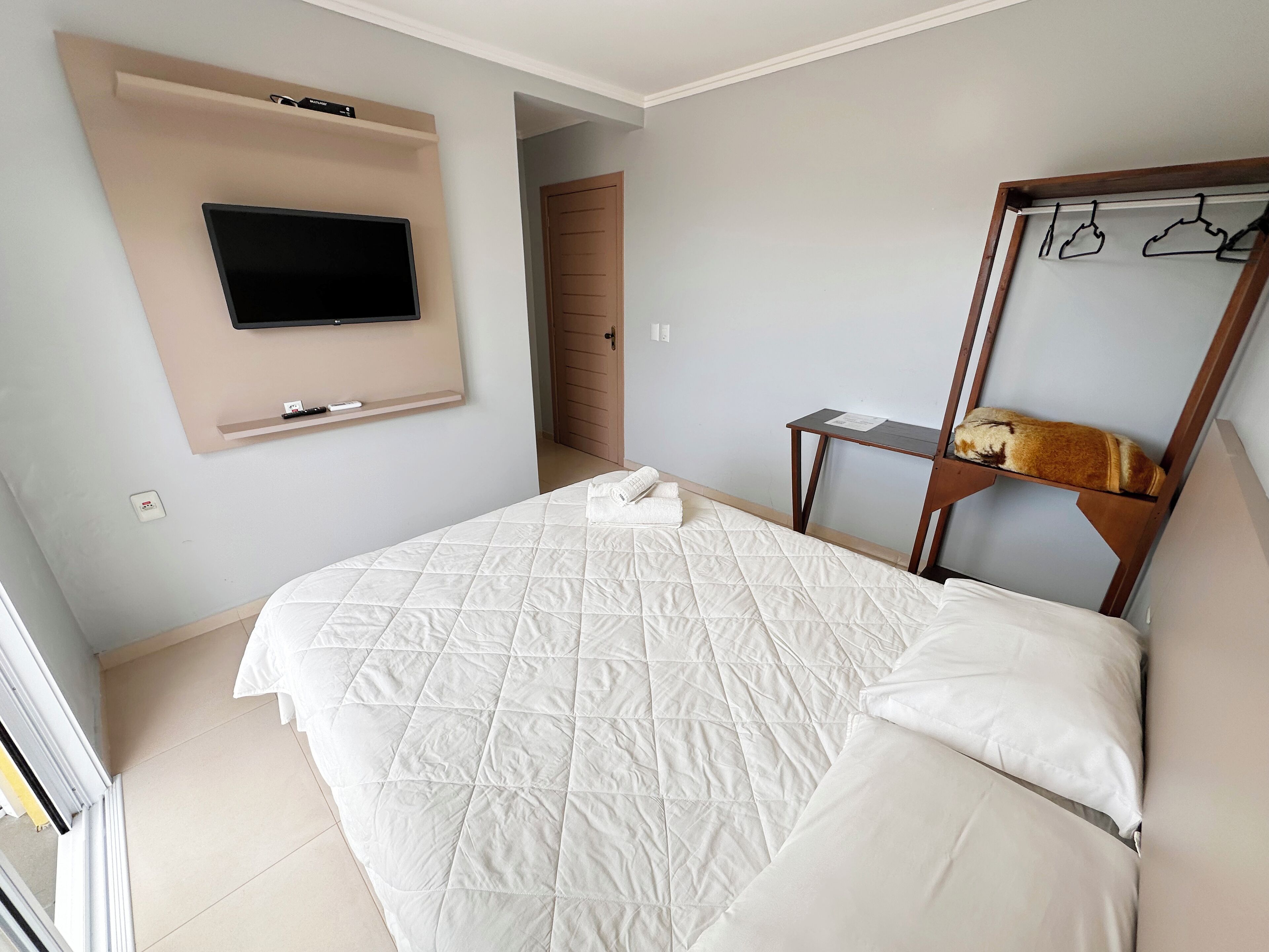 A neatly arranged bedroom with a bed, a mounted TV, a mirror, and a coat rack.