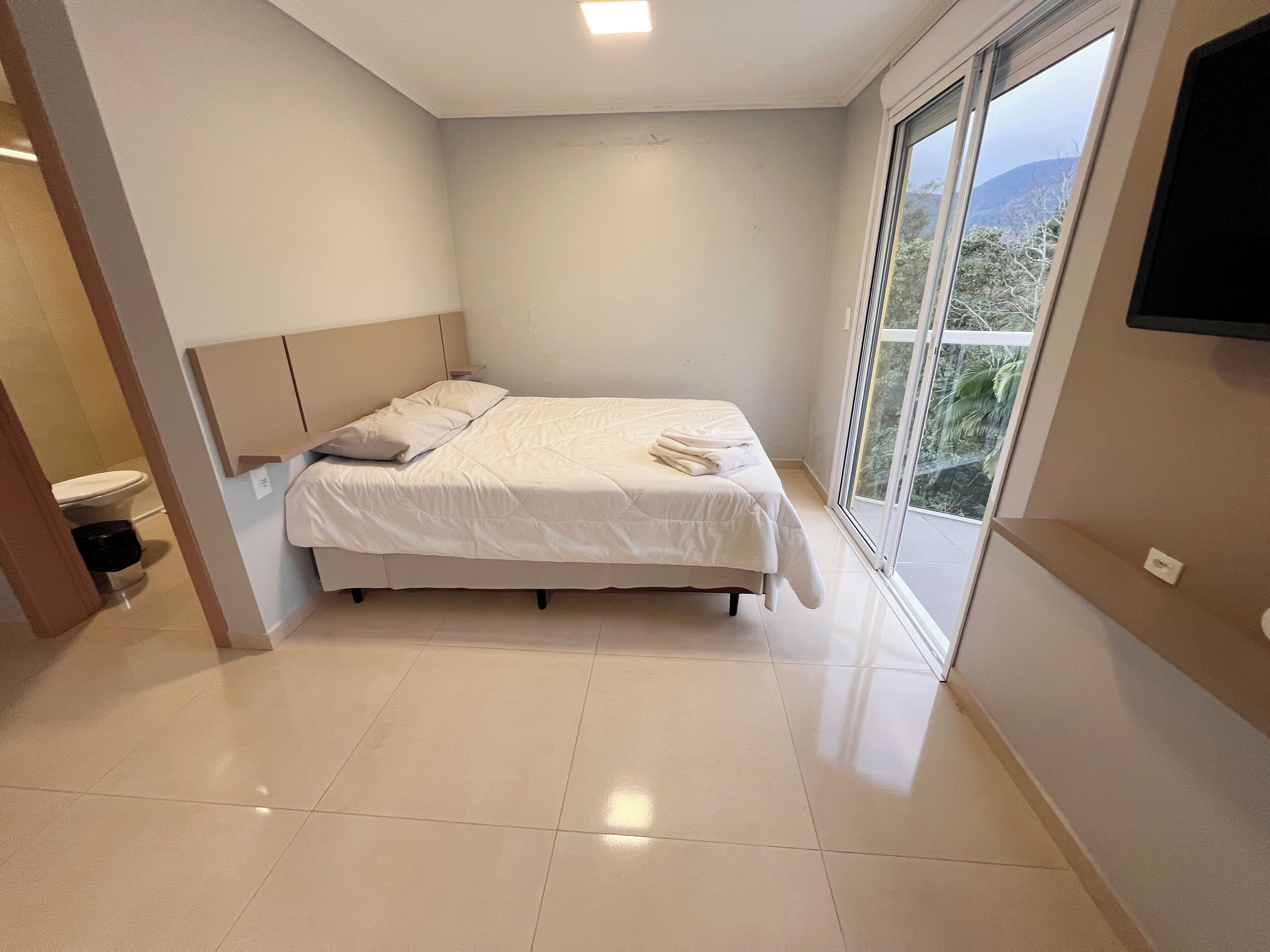 A modern bedroom with a large bed, a window with a view, and a flat-screen TV mounted on the wall.