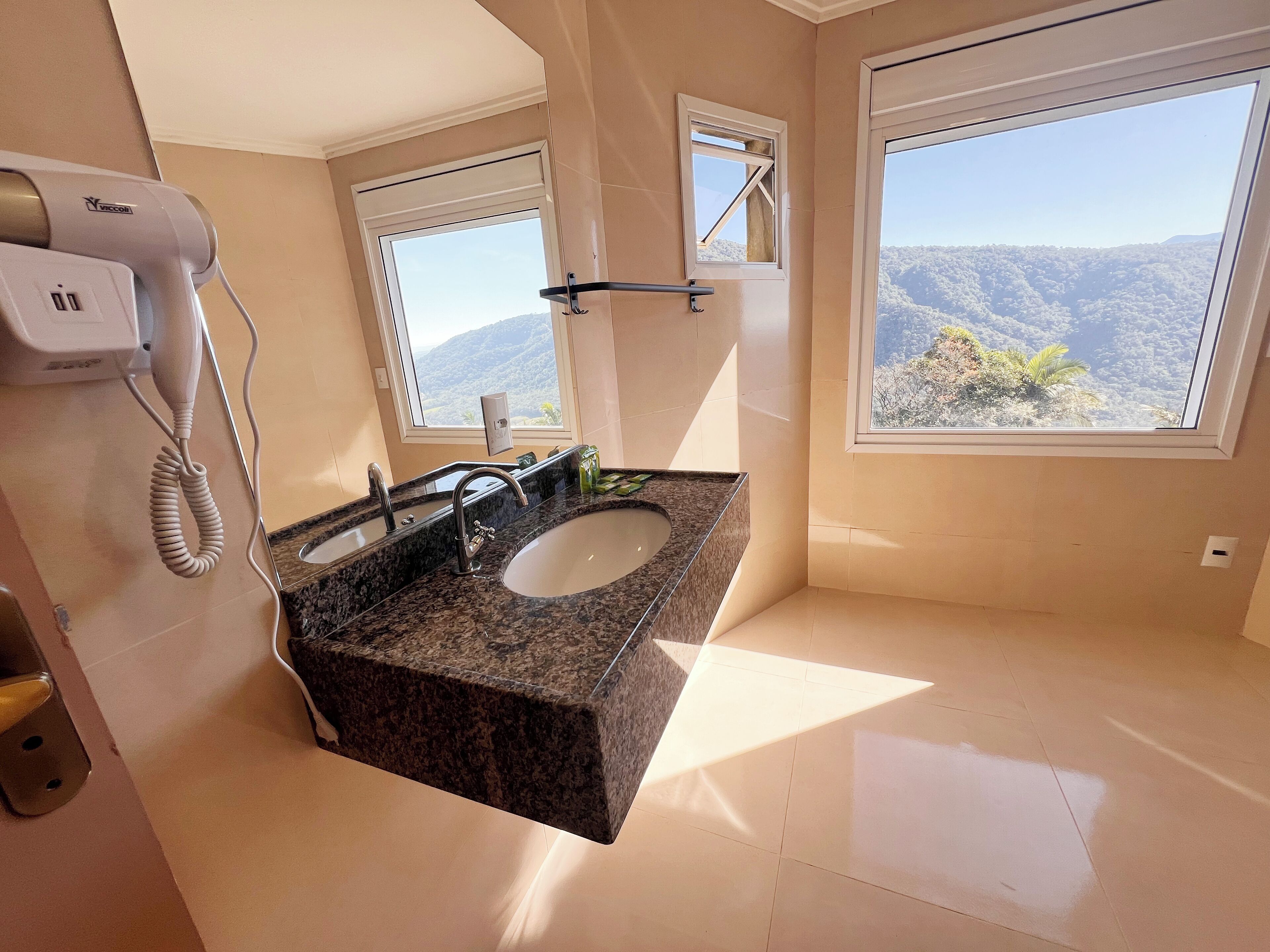 A bathroom with a granite sink, two faucets, a hairdryer, and large windows offering a mountain view.