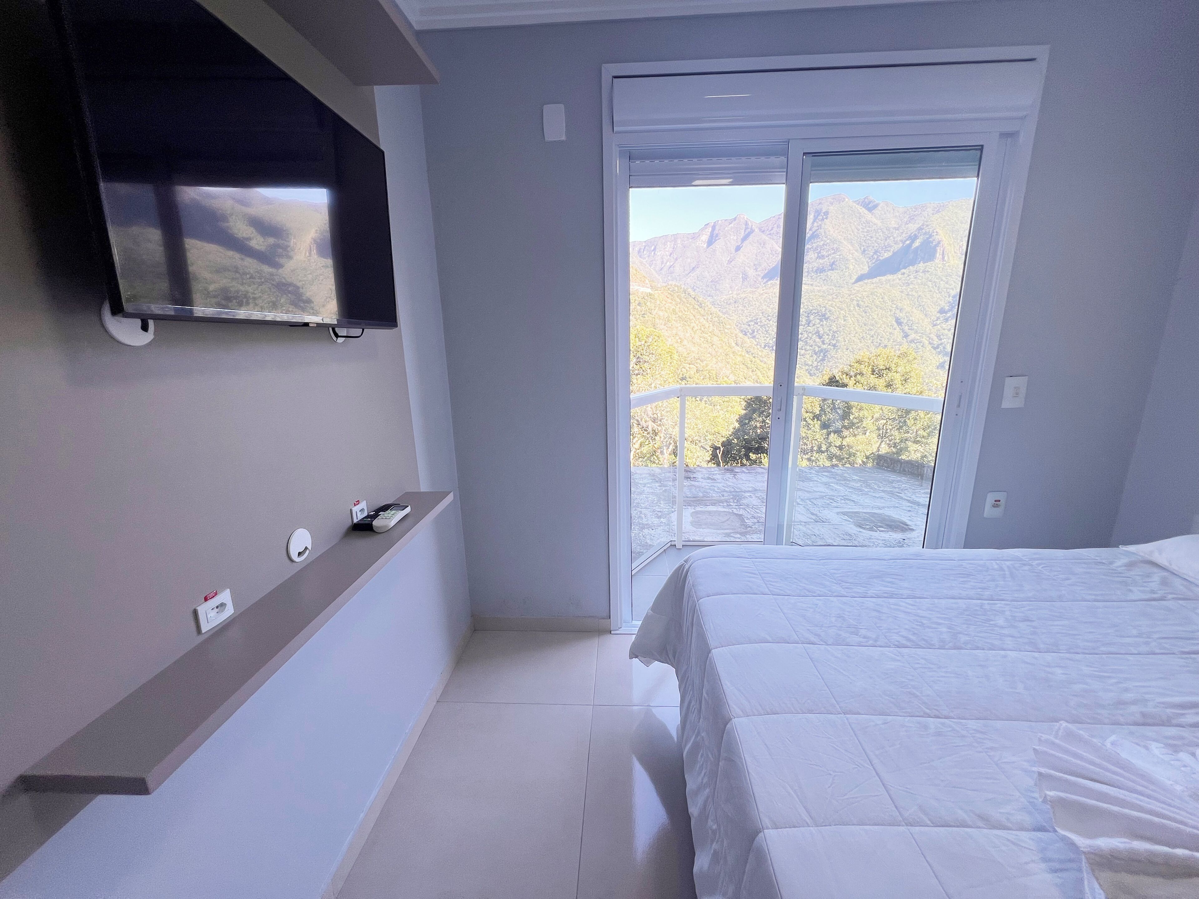 A bedroom with a bed, a TV mounted on the wall, a small shelf with a phone, and a sliding glass door leading to a balcony with a mountain view.