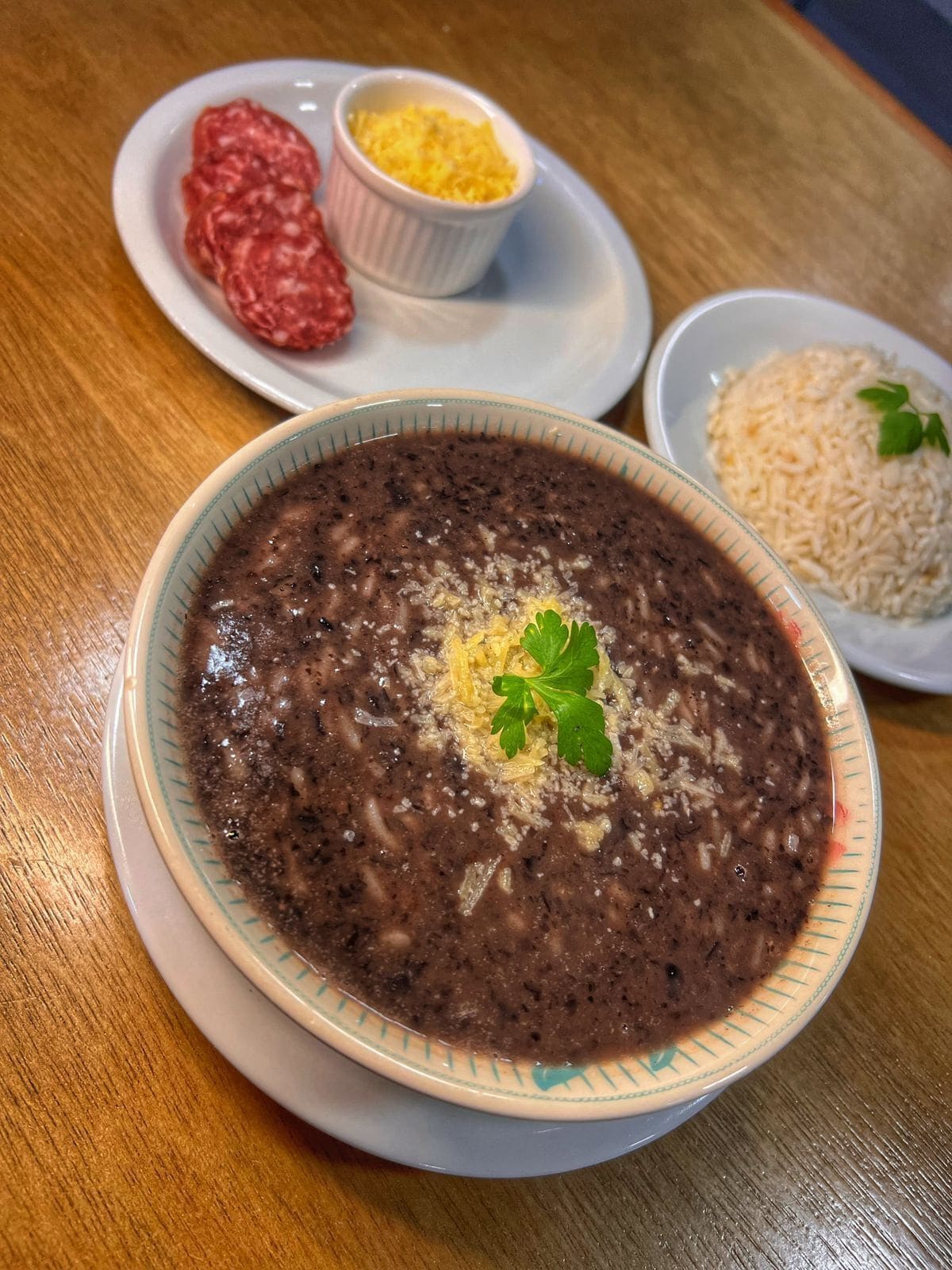 A bowl of black bean soup garnished with cheese and parsley, served with a side of rice and a small dish of meat and cheese.