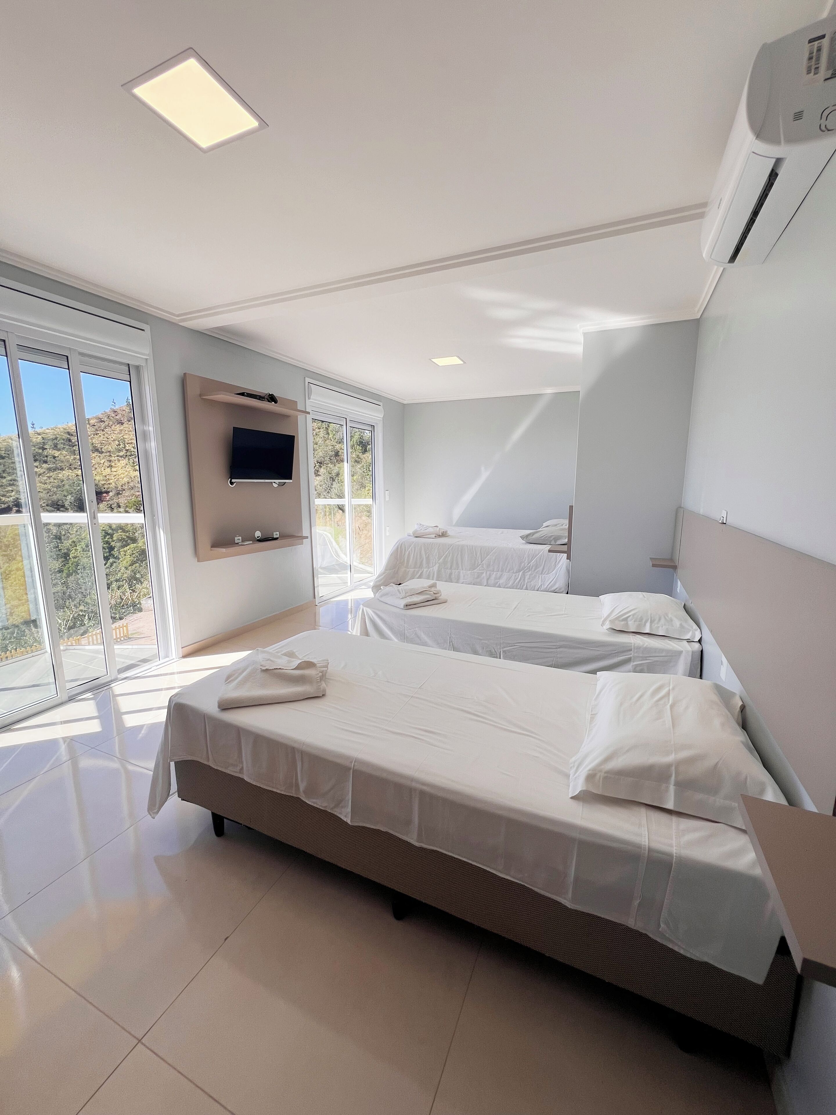 A modern hotel room with two beds, a flat-screen TV, and a large window offering a view of the outdoors.