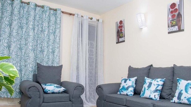 A room with two grey sofas, a large green plant, and a patterned curtain. - Stay.Plus Royale Village Apartment (Syokimau)