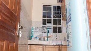 A compact kitchen with wooden cabinets, a white refrigerator, and a window with blinds. - Stay.Plus Royale Village Apartment (Syokimau)