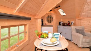 A cozy wooden cabin with a dining area featuring a table with a bowl of fruit, a plate, and a mug. There's a kitchenette with a coffee maker and a cabinet. A striped armchair and a window with a view of the outdoors are also visible.