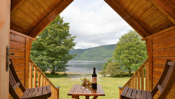 A wooden deck with a table and two chairs, overlooking a lake and hills.