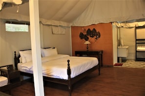 A bedroom with a four-poster bed, a nightstand, a bedside table, a wall-mounted lamp, and a bathroom with a sink and mirror. - The Bugyal (Tehri)