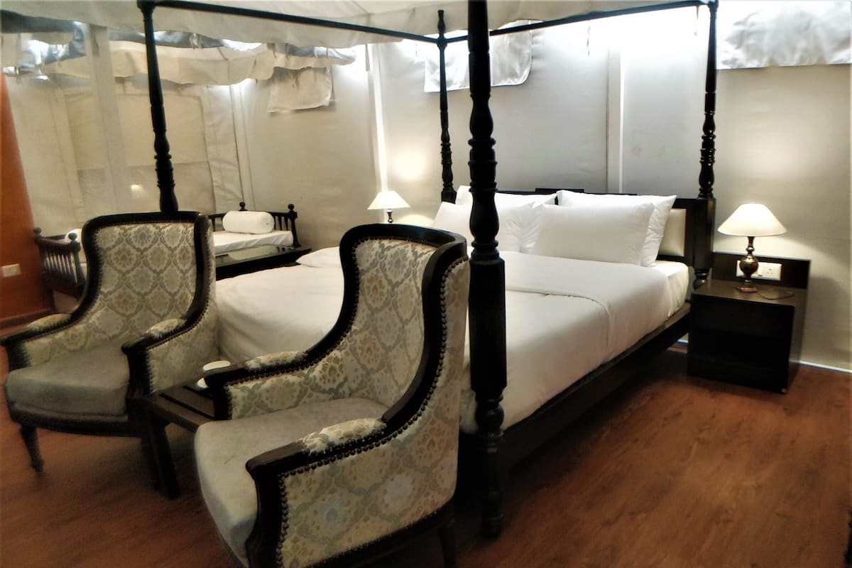 Una cama con dosel y cuatro columnas, con dos sillones tapizados, una mesita de noche y una lámpara.