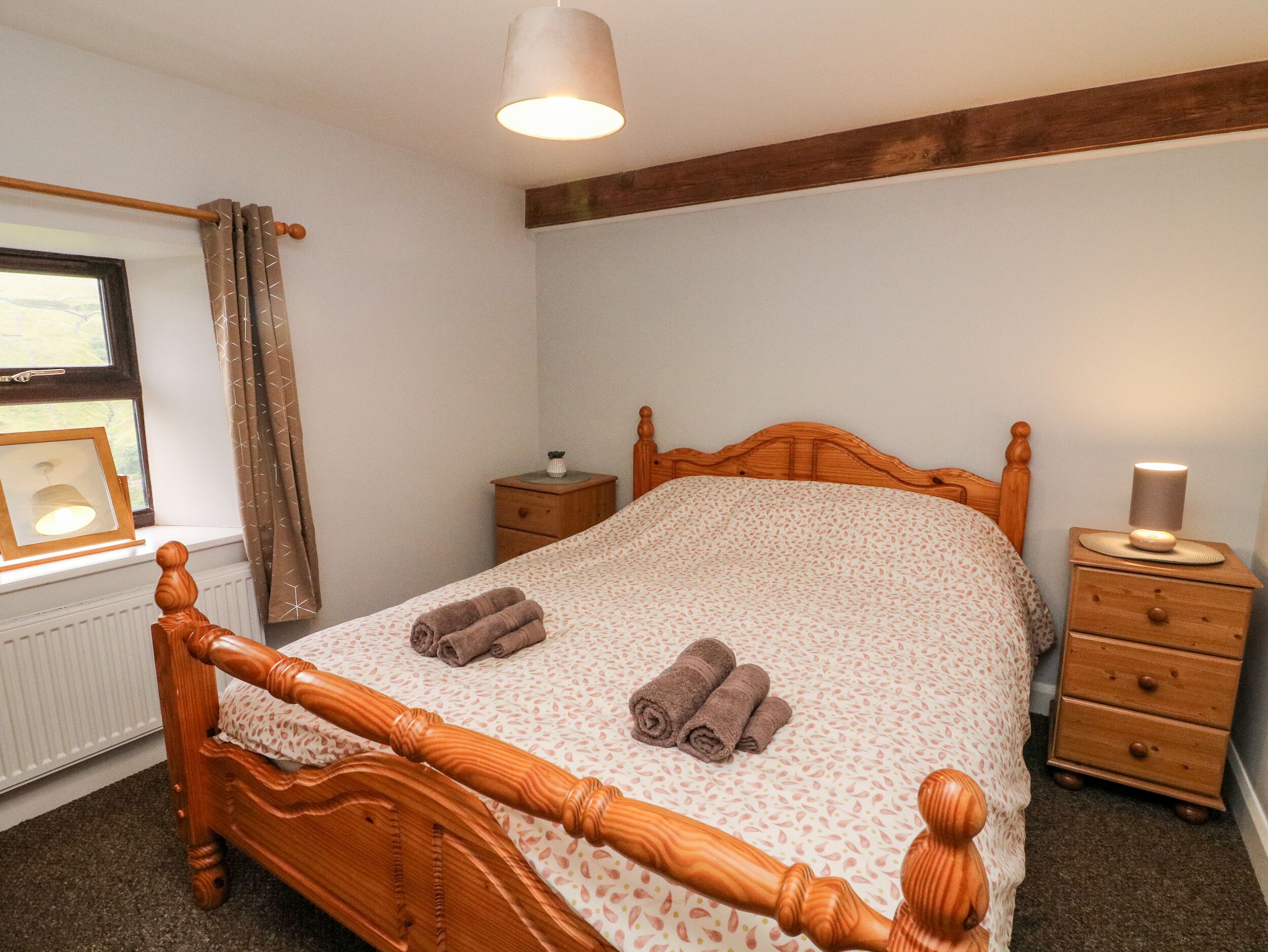 A bedroom with a wooden bed, a bedside table, a nightstand, a lamp, a window with curtains, and a radiator.