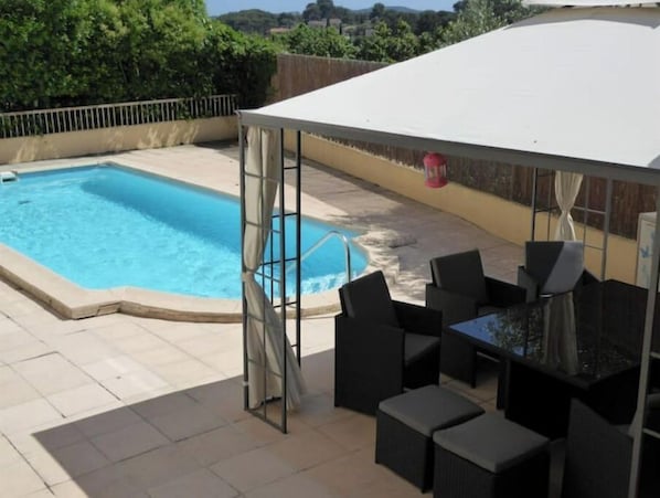 Relax in a shaded area surrounded by lush greenery and a distant hill view, with a pool and comfortable seating.