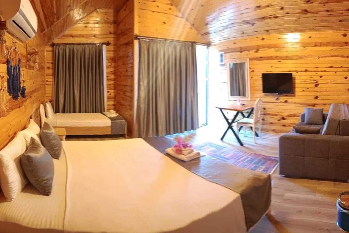 A wooden cabin room with a bed, a sofa, a small table, and a TV mounted on the wall.