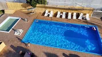 Una piscina rectangular con una cubierta de madera alrededor, una mesa de billar pequeña y varias sillas de descanso.