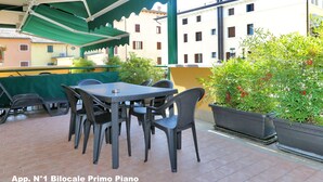 Patio esterno con tavolo e sedie, circondato da piante in vaso e un tenda verde.