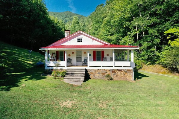 A white house with a red roof, stone foundation, and a porch with a chair. - O'Henry's Getaway, on Cheoah River (Robbinsville)