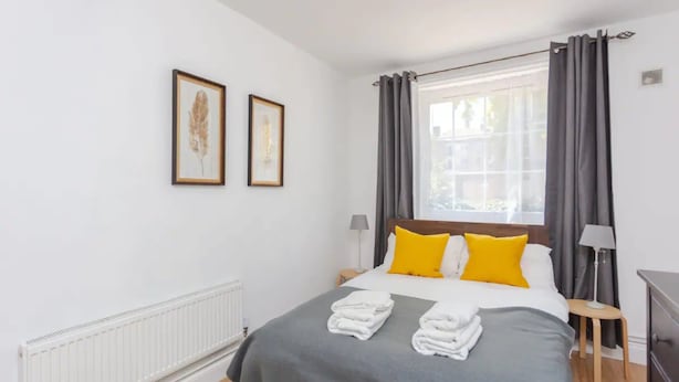 Ein ordentlich bezogenes Bett mit einer grauen Bettdecke und zwei gelben Kissen, ein weißer Heizkörper und zwei gerahmte Bilder an der Wand.