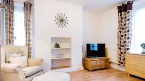 Ein Wohnzimmer mit Sofa, Hocker, Fernseher, Holzschrank, Regalen und einer Wanduhr.