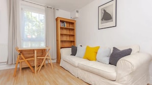 Ein Zimmer mit einem Holztisch, einem weißen Sofa mit Kissen, einem Bücherregal und einem gerahmten Bild an der Wand.