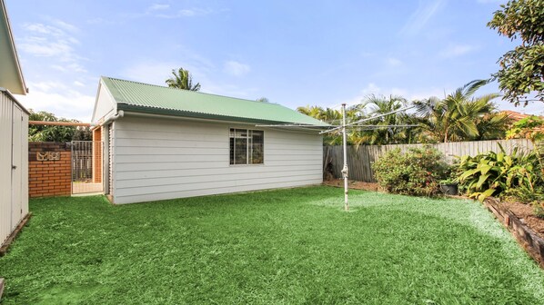A single-story house with a green roof, a fenced backyard, and a brick wall on the left.