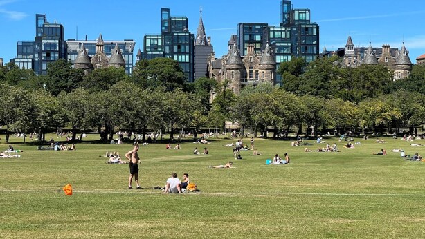 Un parc où des personnes se détendent sur l’herbe, entourées d’arbres et avec des bâtiments historiques en arrière-plan.