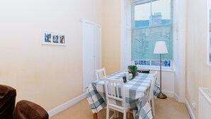 Un coin repas avec une nappe à carreaux, des chaises blanches, une fenêtre avec des stores, une lampe sur pied et une petite plante.