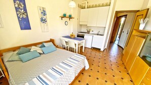 Un dormitorio con cama, una mesita pequeña y sillas. Se observa una zona de cocina con armarios y un lavamanos.