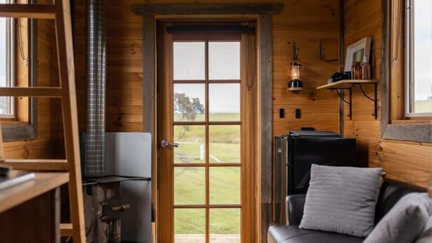 A cozy cabin interior with a wooden door leading to a balcony, a fireplace, a sofa, and a shelf with decorative items.