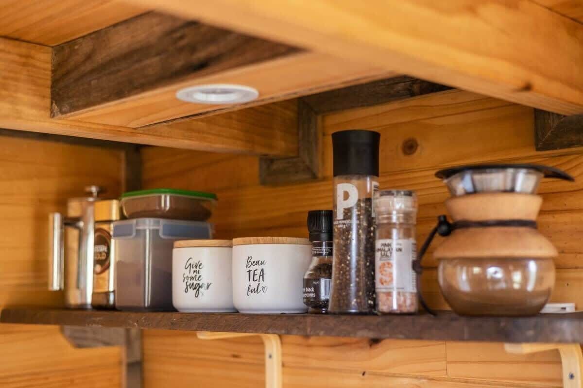 Ein Holzregal mit verschiedenen Behältern und Küchenutensilien, darunter Gläser mit den Aufschriften „Gib Zucker“ und „Tee anbieten“.