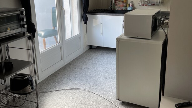 A compact kitchen with white cabinets, a small refrigerator, a toaster, and a window with blinds. - Appartement MTM (Montpellier)