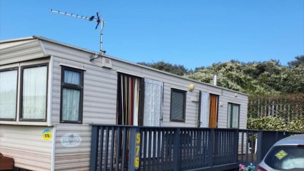 A mobile home with a fenced deck, a satellite dish, and a car parked nearby.