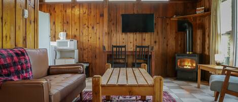 A cozy cabin interior with a wooden interior, a sofa, a dining table, chairs, a TV mounted on the wall, and a wood-burning stove.